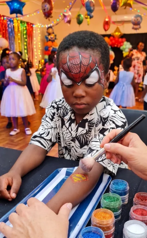 Boy with Spider-Man face paint getting a glitter Superman tattoo on his arm