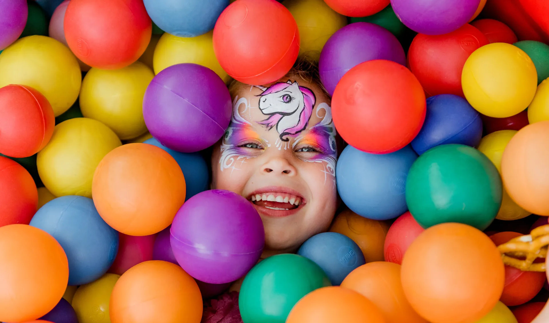 Laughing girl with unicorn face paint lying in a colourful ball pit